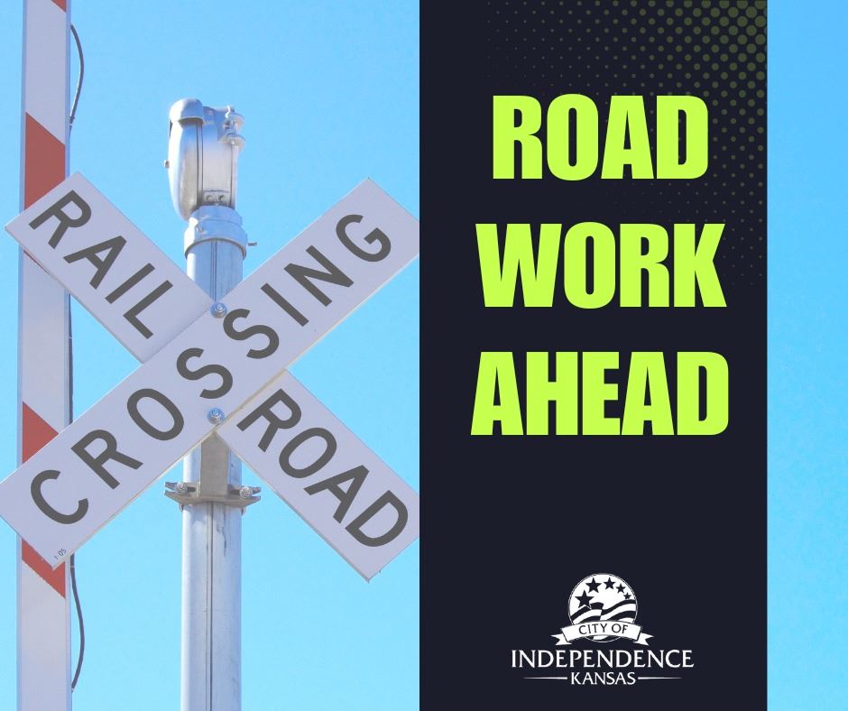 Railroad crossing sign with blue sky; beside it, black banner reads 'ROAD WORK AHEAD'